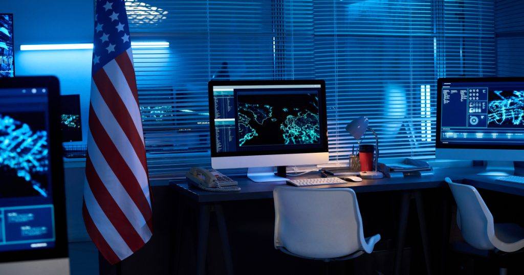 Cybersecurity operations center with U.S. flag and real-time global threat analysis on multiple monitors.
