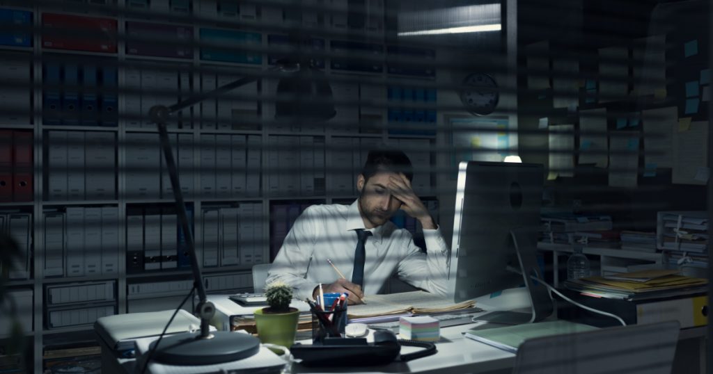A tired business professional working late at night, head in hand, worried about unseen cybersecurity threats.